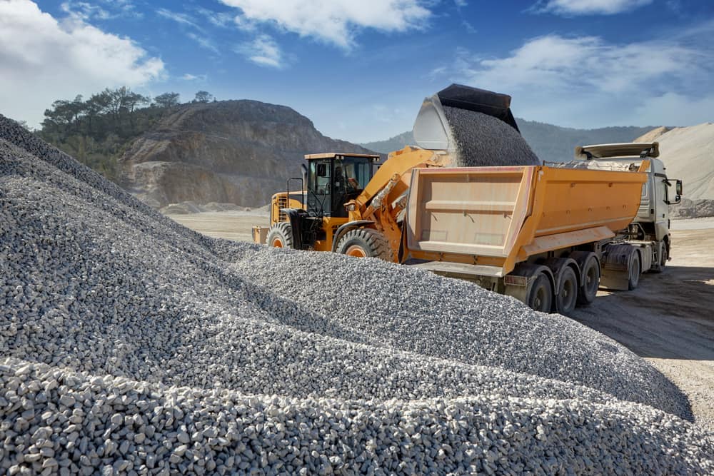 A yellow loader filling an orange dump trailer with gravel at a quarry